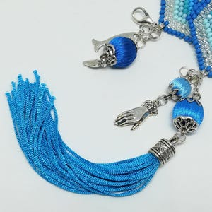 May include: A close-up of a blue beaded bracelet with silver-toned charms and a long blue tassel. The charms include a whale, a hand, and blue silk-covered beads. The bracelet has a clasp closure.