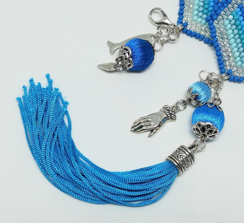 May include: A close-up of a blue beaded bracelet with silver-toned charms and a long blue tassel. The charms include a whale, a hand, and blue silk-covered beads. The bracelet has a clasp closure.