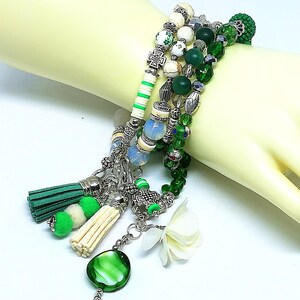 May include: A multi-strand bracelet featuring green, white, and silver beads. The bracelet includes decorative tassels, a green glass pendant, and a white flower charm. The beads vary in size and shape, creating a layered effect.