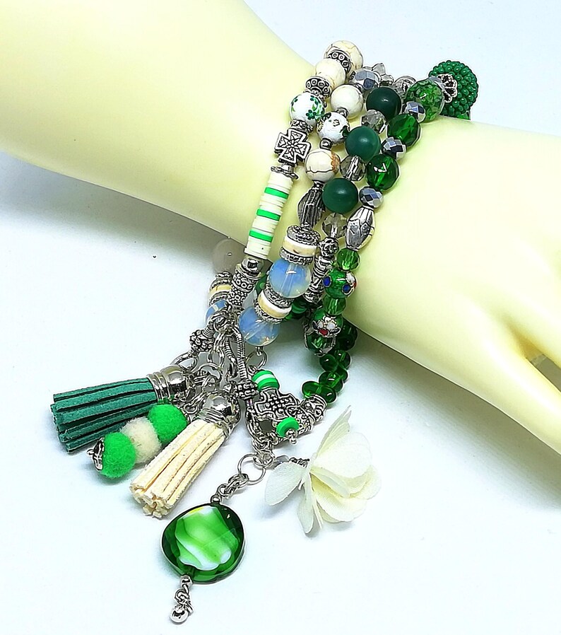 May include: A multi-strand bracelet featuring green, white, and silver beads. The bracelet includes decorative tassels, a green glass pendant, and a white flower charm. The beads vary in size and shape, creating a layered effect.