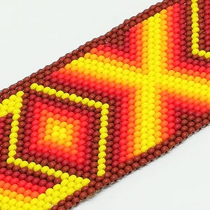 Op de afbeelding: Close-up van een kralenarmband met een geometrisch ontwerp. De armband heeft een patroon van gele, oranje en rode kralen, met een bruine rand. De kralen zijn klein en dicht op elkaar gepakt, waardoor een textuur ontstaat.