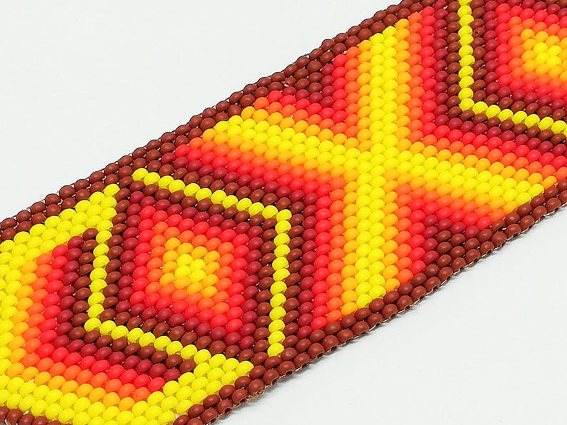 Op de afbeelding: Close-up van een kralenarmband met een geometrisch ontwerp. De armband heeft een patroon van gele, oranje en rode kralen, met een bruine rand. De kralen zijn klein en dicht op elkaar gepakt, waardoor een textuur ontstaat.