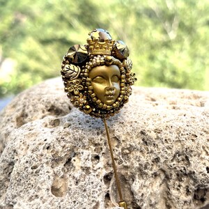 May include: A gold-toned hat pin featuring a detailed face with a crown, surrounded by decorative elements. The pin has a long, slender shaft and is set against a blurred green background.