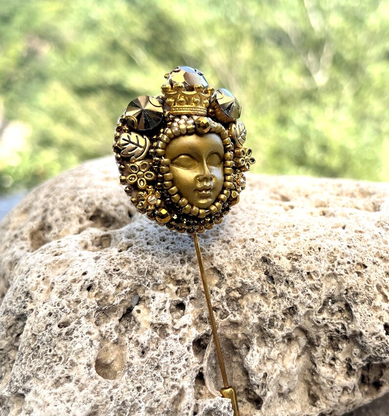 May include: A gold-toned hat pin featuring a detailed face with a crown, surrounded by decorative elements. The pin has a long, slender shaft and is set against a blurred green background.