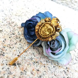 May include: A gold-toned lapel pin featuring a detailed face design, adorned with a crown and surrounded by small beads. The pin is set against a backdrop of blue and light blue artificial roses, creating a decorative accessory.