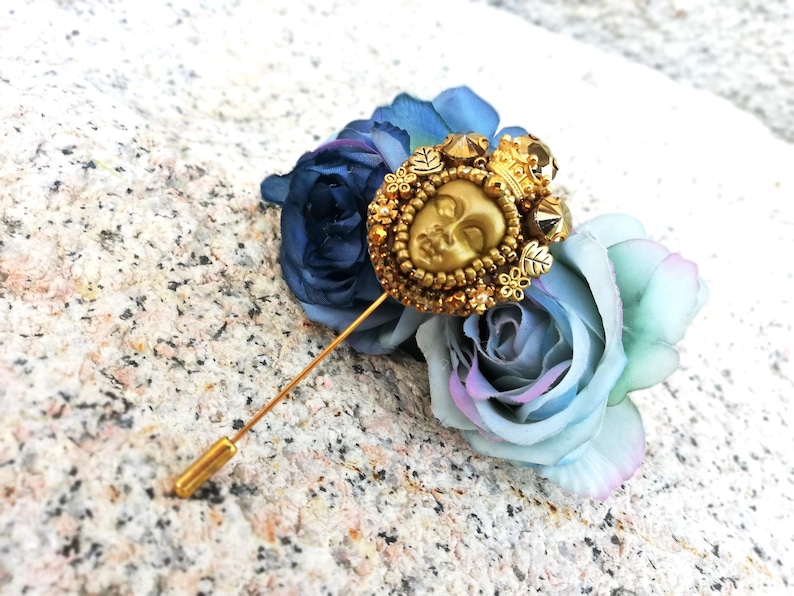 May include: A gold-toned lapel pin featuring a detailed face design, adorned with a crown and surrounded by small beads. The pin is set against a backdrop of blue and light blue artificial roses, creating a decorative accessory.