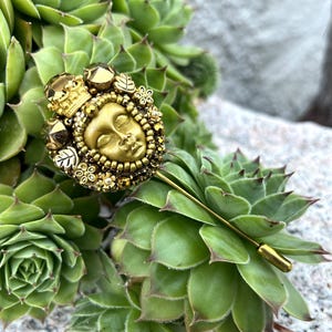 May include: A gold-toned stick pin featuring a detailed face design, surrounded by floral and geometric embellishments, and topped with a small crown. The pin is set against a backdrop of green succulent plants.