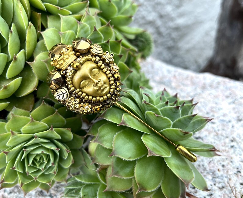 May include: A gold-toned stick pin featuring a detailed face design, surrounded by floral and geometric embellishments, and topped with a small crown. The pin is set against a backdrop of green succulent plants.