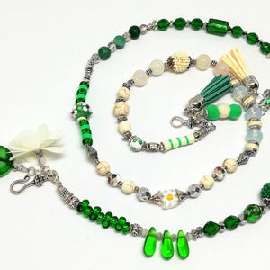 May include: A collection of handmade beaded jewelry in shades of green, white, and silver. The pieces include necklaces, bracelets, and decorative tassels. The beads vary in size and shape, with some featuring floral and geometric designs.