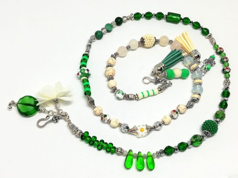 May include: A collection of handmade beaded jewelry in shades of green, white, and silver. The pieces include necklaces, bracelets, and decorative tassels. The beads vary in size and shape, with some featuring floral and geometric designs.