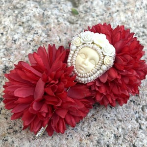 May include: A white porcelain face with closed eyes surrounded by white flowers and pearls, attached to a red fabric flower hair clip.