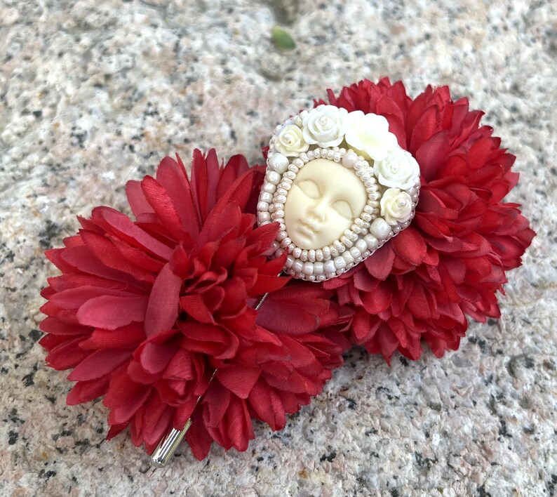 May include: A white porcelain face with closed eyes surrounded by white flowers and pearls, attached to a red fabric flower hair clip.