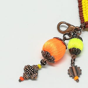 Op de afbeelding: Close-up van een handgemaakte armband met een koperen sluiting en decoratieve bedels. De armband heeft oranje en gele kralen, met oranje en gele zijde omwikkelde kralen als bedels. Extra bedels zijn een tandwiel en een decoratief vierkant.