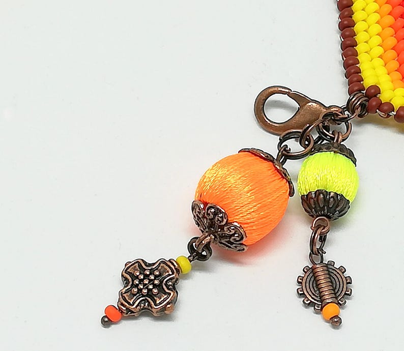 Op de afbeelding: Close-up van een handgemaakte armband met een koperen sluiting en decoratieve bedels. De armband heeft oranje en gele kralen, met oranje en gele zijde omwikkelde kralen als bedels. Extra bedels zijn een tandwiel en een decoratief vierkant.