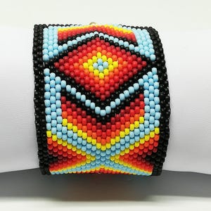 May include: A wide beaded bracelet featuring a geometric pattern in shades of red, orange, yellow, blue, and black. The bracelet is made of small, colorful beads and is displayed on a white surface.