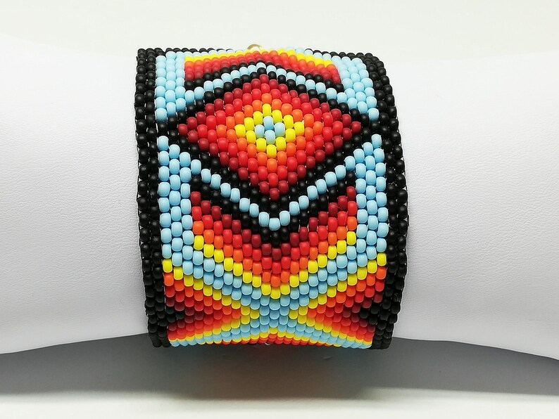 May include: A wide beaded bracelet featuring a geometric pattern in shades of red, orange, yellow, blue, and black. The bracelet is made of small, colorful beads and is displayed on a white surface.