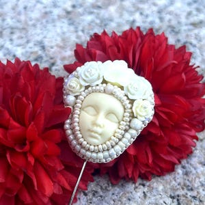 May include: A white porcelain face with closed eyes surrounded by white flowers and white beads, on a red flower background. The face is attached to a silver pin.