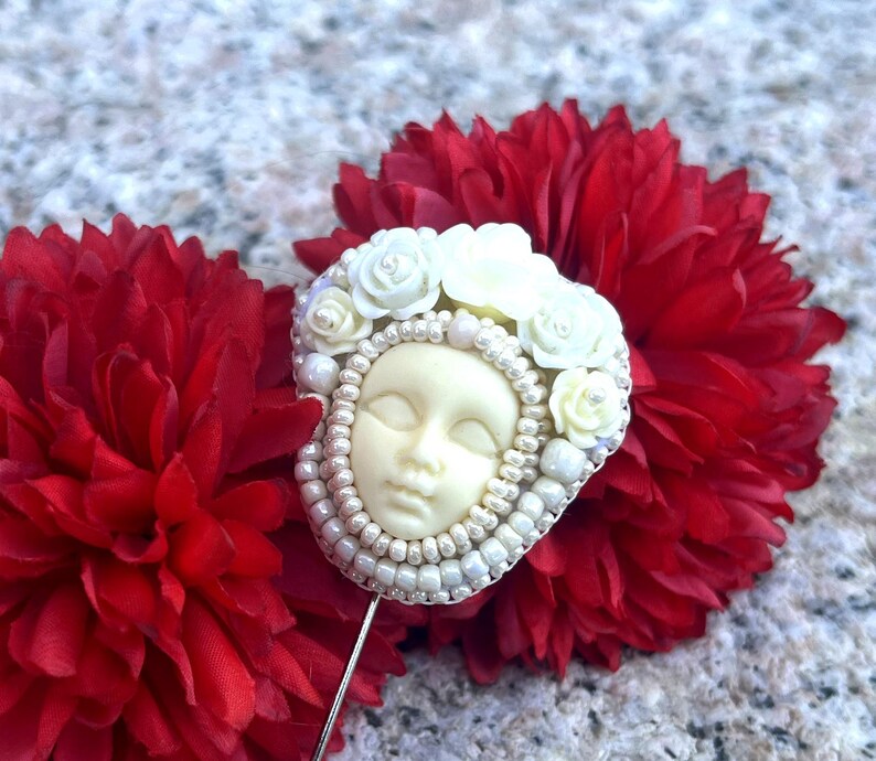 May include: A white porcelain face with closed eyes surrounded by white flowers and white beads, on a red flower background. The face is attached to a silver pin.