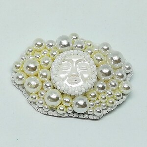 May include: A white pearl brooch with a white face in the center. The face has closed eyes and a slightly open mouth. The brooch is surrounded by a cluster of white pearls.