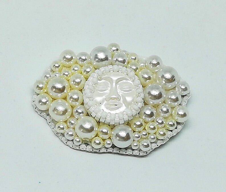 May include: A white pearl brooch with a white face in the center. The face has closed eyes and a slightly open mouth. The brooch is surrounded by a cluster of white pearls.