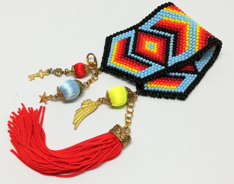 May include: A beaded cuff bracelet with a geometric pattern in red, yellow, blue, and black. The bracelet has gold-colored charms, including a key, star, and wing, and a red tassel. The bracelet is approximately 2 inches wide.