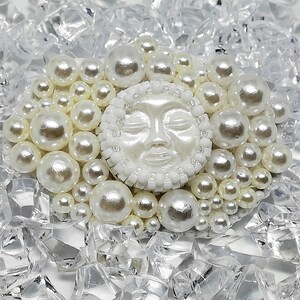May include: A white ceramic sun face with closed eyes, surrounded by a ring of white pearls, set against a background of clear, broken glass.