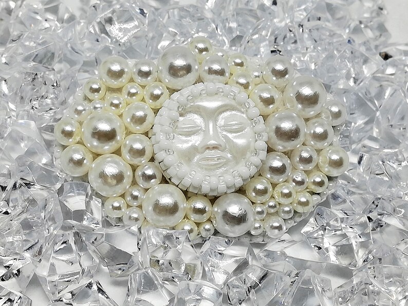 May include: A white ceramic sun face with closed eyes, surrounded by a ring of white pearls, set against a background of clear, broken glass.