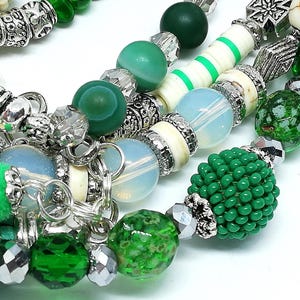 May include: Close-up of beaded necklaces with a variety of green and silver beads. The necklaces feature different textures, including smooth, faceted, and beaded elements. Some beads are a light blue color.