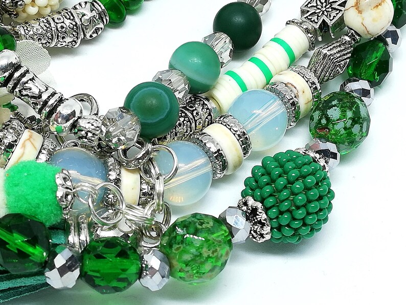 May include: Close-up of beaded necklaces with a variety of green and silver beads. The necklaces feature different textures, including smooth, faceted, and beaded elements. Some beads are a light blue color.