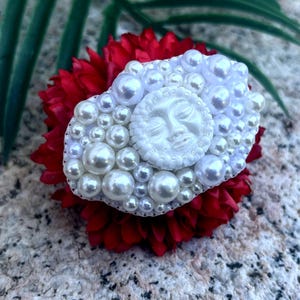 May include: A white pearl and resin brooch with a sun face design. The brooch is set on a red flower.
