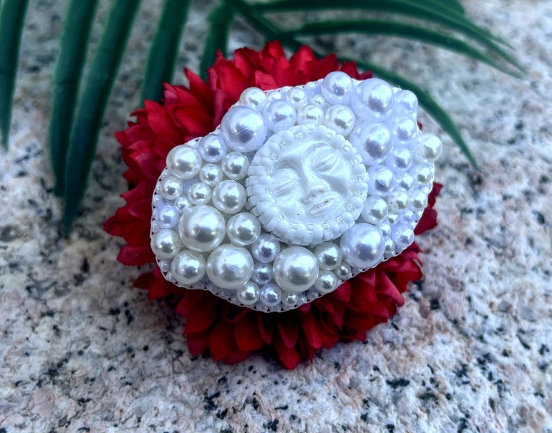 May include: A white pearl and resin brooch with a sun face design. The brooch is set on a red flower.