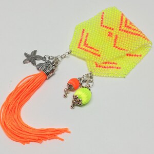 May include: A yellow beaded accessory with orange detailing. It features a bright orange tassel, a starfish charm, and two beaded orbs in orange and green. The accessory has a geometric design in orange.