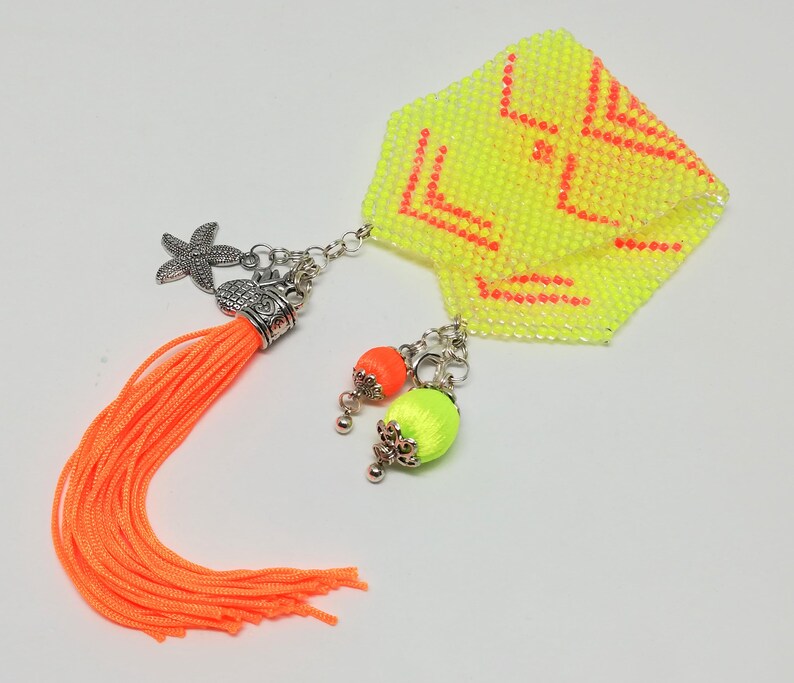 May include: A yellow beaded accessory with orange detailing. It features a bright orange tassel, a starfish charm, and two beaded orbs in orange and green. The accessory has a geometric design in orange.