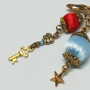 May include: A gold-toned charm bracelet with decorative elements. Features a key, leaf, star, and two orb-shaped charms, one red and one light blue. The charms are connected by gold chains and clasps.
