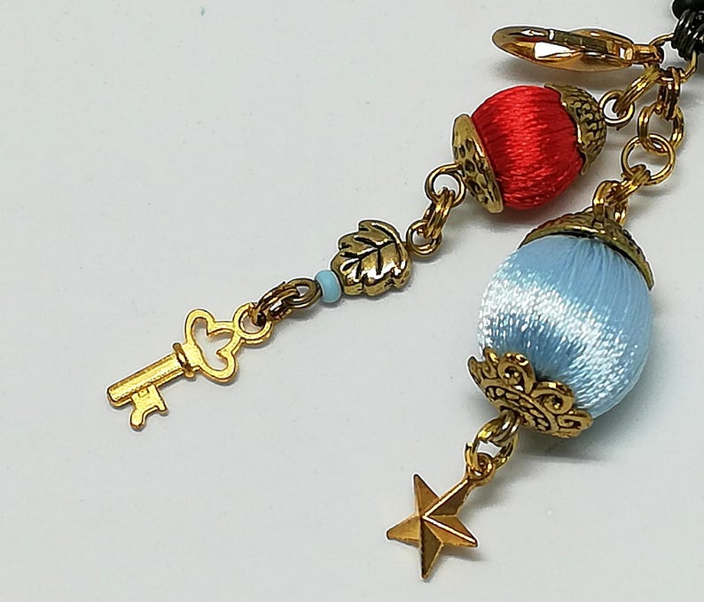 May include: A gold-toned charm bracelet with decorative elements. Features a key, leaf, star, and two orb-shaped charms, one red and one light blue. The charms are connected by gold chains and clasps.