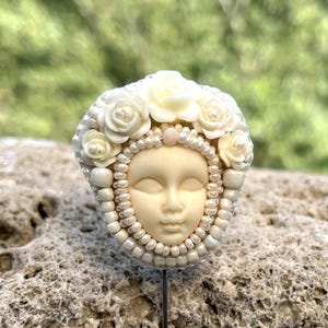 May include: A white porcelain brooch with a face and a crown of white flowers and pearls.