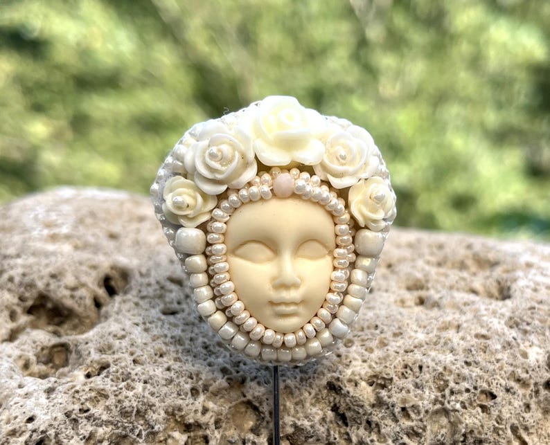 May include: A white porcelain brooch with a face and a crown of white flowers and pearls.