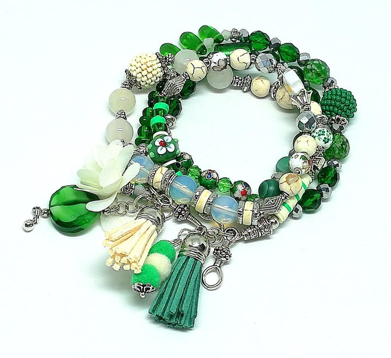 May include: A collection of beaded bracelets in shades of green, white, and silver. The bracelets feature various bead shapes and sizes, including round, faceted, and textured beads. Decorative tassels and charms add detail to the bracelets. A white flower and green glass bead are also included.