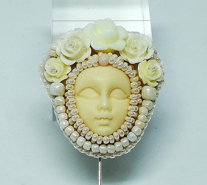 May include: A white porcelain face with closed eyes and a delicate smile, surrounded by a wreath of white flowers and white seed beads.