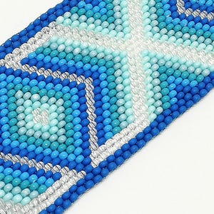 May include: Close-up of a beaded bracelet with a geometric pattern. The bracelet features a variety of blue and turquoise beads, with silver and white accents. The design is a repeating pattern of squares and diamonds.