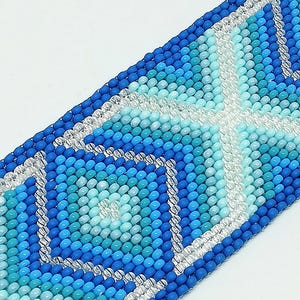 May include: A beaded bracelet in shades of blue and clear beads. The bracelet features a geometric pattern with a central cross design. The beads are small and round, creating a textured surface. The bracelet is likely handmade.