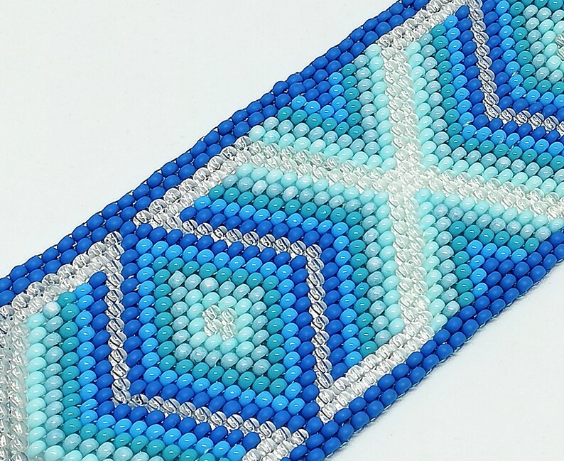 May include: A beaded bracelet in shades of blue and clear beads. The bracelet features a geometric pattern with a central cross design. The beads are small and round, creating a textured surface. The bracelet is likely handmade.