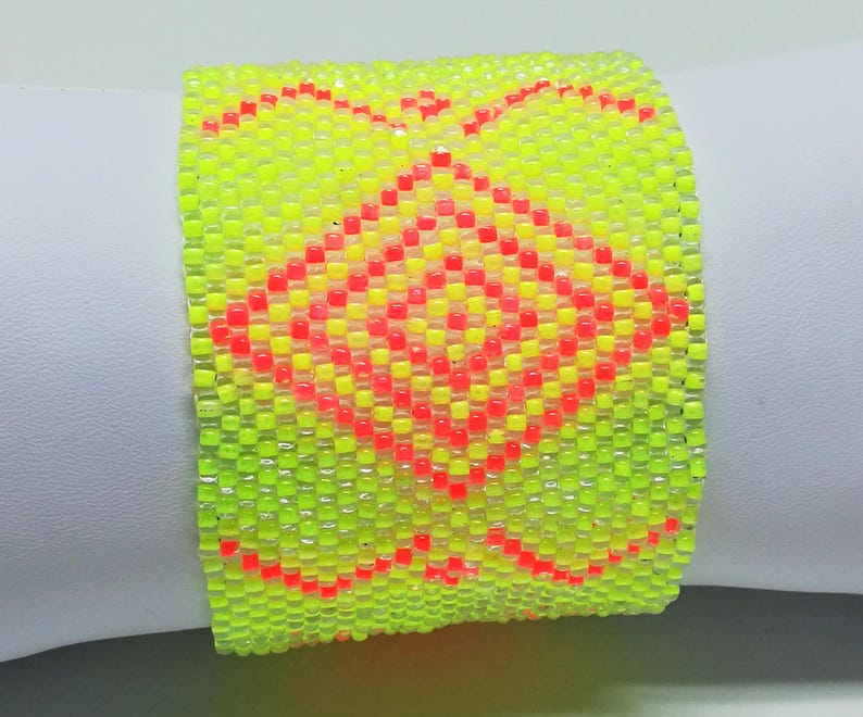 May include: A neon yellow beaded bracelet with a geometric pattern in red and orange. The bracelet is wide and appears to be made of small, square beads. The design features a central diamond shape and decorative edges.