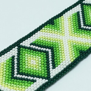 May include: Close-up of a beaded bracelet with a geometric pattern. The bracelet features a repeating design of white, light green, and dark green beads. The beads are arranged in a diamond and square pattern. The bracelet is on a white background.