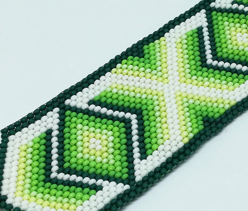 May include: Close-up of a beaded bracelet with a geometric pattern. The bracelet features a repeating design of white, light green, and dark green beads. The beads are arranged in a diamond and square pattern. The bracelet is on a white background.