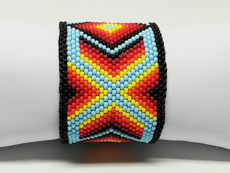 May include: A wide beaded bracelet with a colorful geometric pattern. The bracelet features a black border and a central design in red, orange, yellow, and light blue. The bracelet is displayed on a white surface.