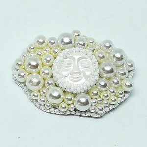 May include: A white pearl brooch with a white face in the center. The face has closed eyes and a slightly open mouth. The brooch is surrounded by white pearls of varying sizes.