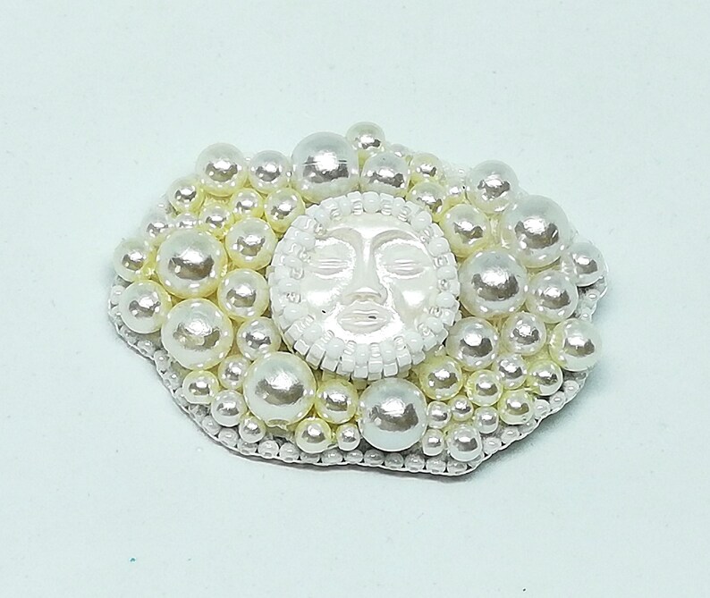 May include: A white pearl brooch with a white face in the center. The face has closed eyes and a slightly open mouth. The brooch is surrounded by white pearls of varying sizes.