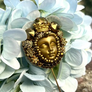 May include: A gold-toned decorative pin featuring a serene face with a crown, surrounded by intricate beadwork and embellishments. The pin is set against a backdrop of light blue hydrangea flowers, creating a striking contrast.
