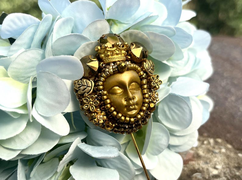 May include: A gold-toned decorative pin featuring a serene face with a crown, surrounded by intricate beadwork and embellishments. The pin is set against a backdrop of light blue hydrangea flowers, creating a striking contrast.